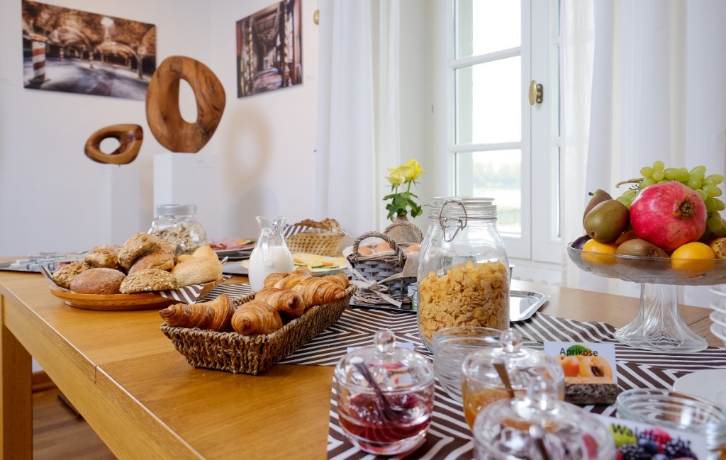 A breakfast table with rolls, croissants, cereal, milk, eggs, jam and fruit.
