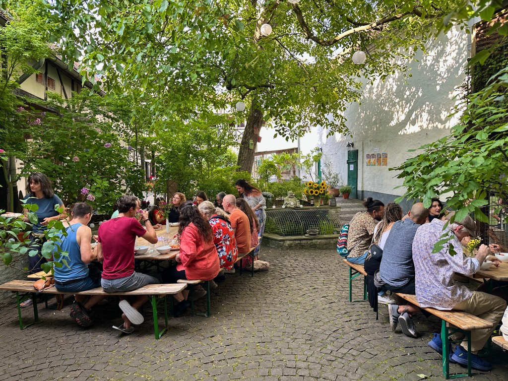 People eating at long tables in the courtyard, under the trees.