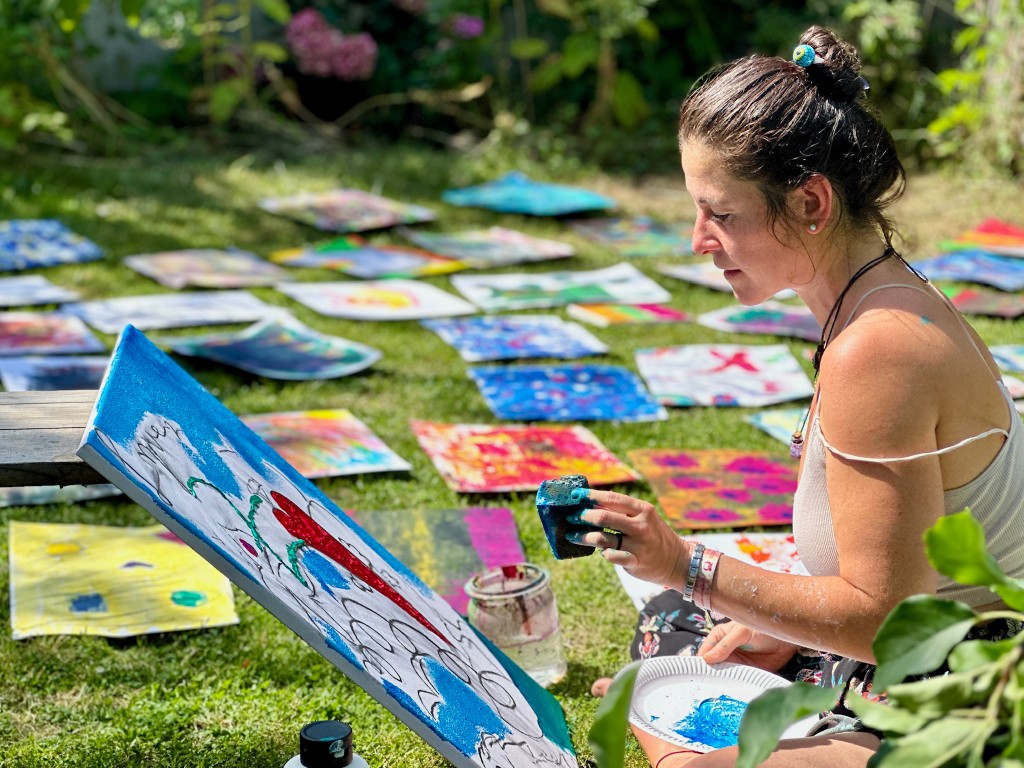 A woman painting, with many paintings laid out on the grass.
