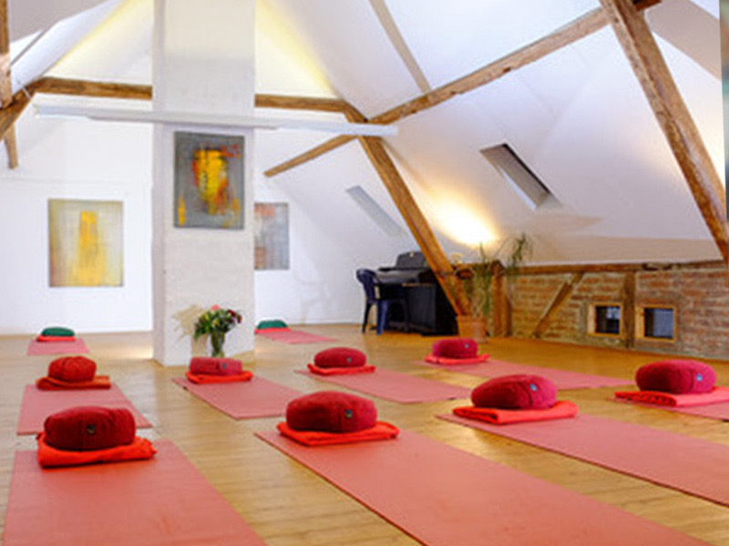 A light room under the roof, with mats laid out, wooden beams, and a piano against the far wall.