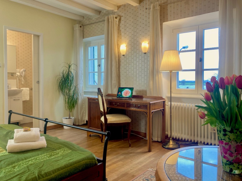 A room with a green bedspread, a writing desk, a lamp, and a view into the bathroom.
