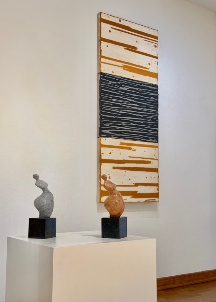 Two small sculptures in front of a tall painting in the gallery.