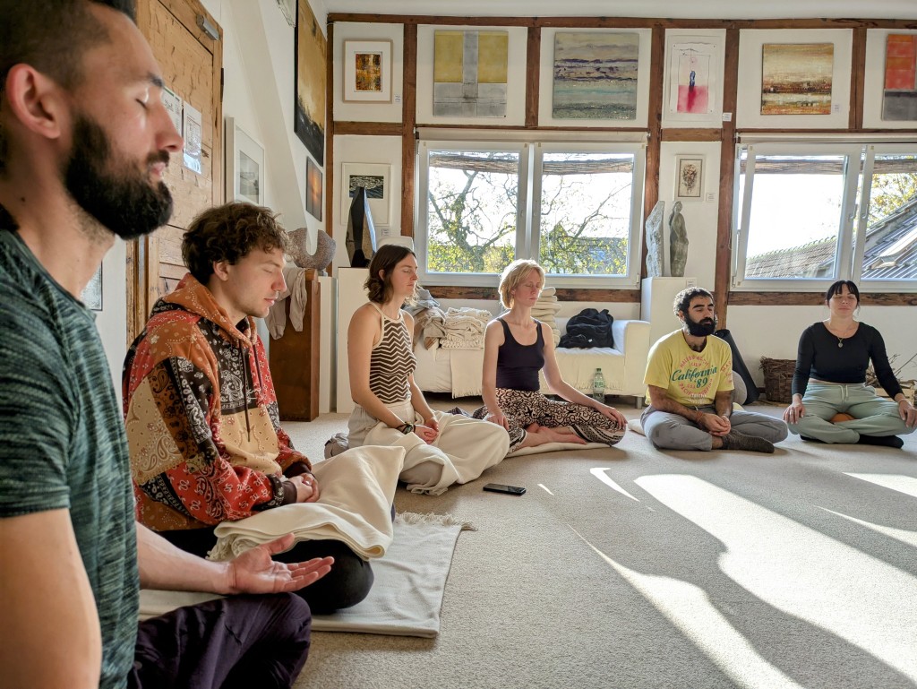 Sitzen im Licht der Fenster. Meditation im hellen Raum des Yoga Hotels am Rhein, mit Bildern an der Wand.